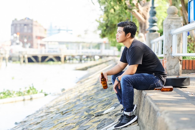 Asiatischer junger Mann, der Bier trinkt und den Blick auf den Fluss betrachtet, den er an seinen Wochenenden im Freien genießt