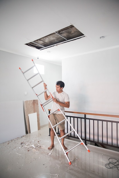 Asiatischer junger Mann, der eine Leiter benutzt, um die gebrochene Decke zu Hause zu reparieren