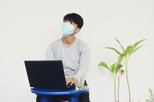 Asiatischer junger Mann, der eine Maske trägt und vor einem Laptop sitzt, der von seinem Job schwindlig ist?