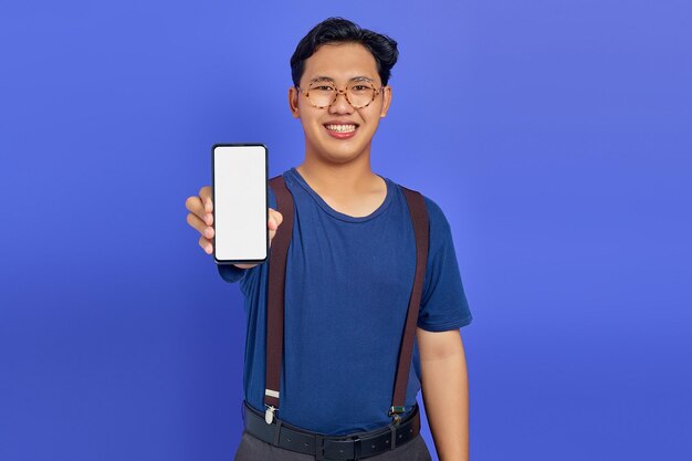 Asiatischer junger Mann, der einen leeren Smartphone-Bildschirm mit der Hand zur Kamera auf violettem Hintergrund zeigt