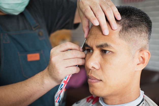 Asiatischer junger Mann, der Haarschnitt mit Rasiermesser durch professionellen Friseur im Friseursalon ist.