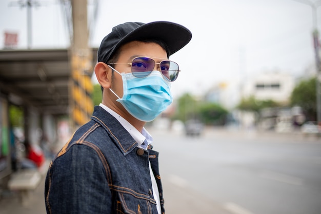 Asiatischer junger Mann, der in der Stadt steht und Schutzmaske auf Gesicht für Schutzluftverschmutzung, Partikel und für Schutzgrippevirus, Influenza, Coronavirus in der Stadt trägt