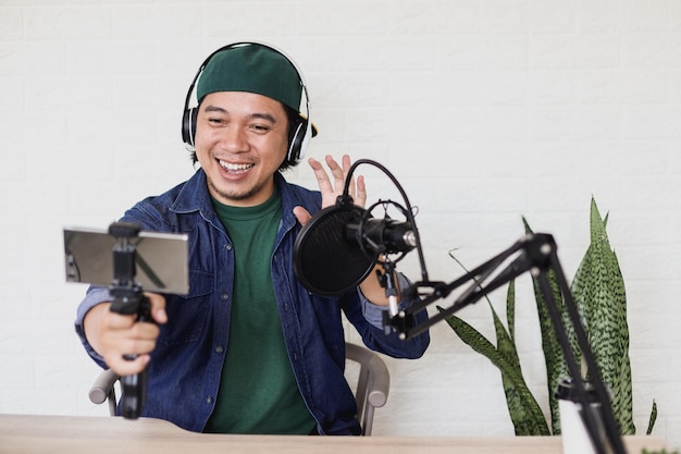 Asiatischer junger mann im lässigen stil macht einen vlog mit dem smartphone im podcasting-raum und sagt hallo