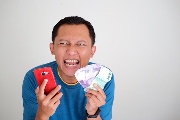 Asiatischer junger Mann mit glücklichem Gesichtsausdruck, während er viel Geld auf grauem Hintergrund isoliert hält