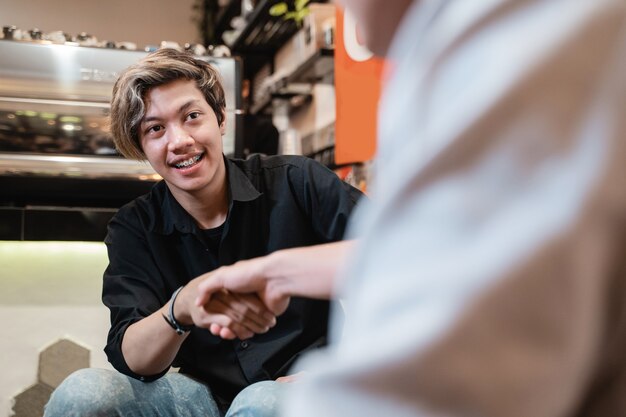 Asiatischer junger Mann schüttelt Hand mit Partner, wenn auf Sofa im Kaffeehaus sitzen