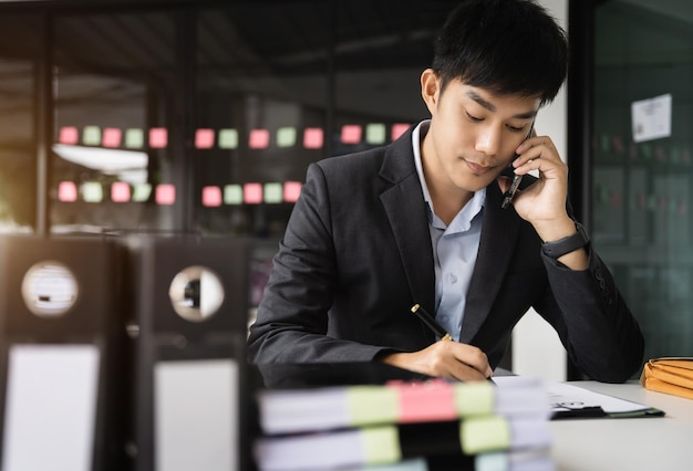 Asiatischer junger Mann spricht mit Mobiltelefonen und macht sich Notizen auf seinen Notizbüchern, während er an Laptop-Computern arbeitet. Online-Business-Telearbeitskonzept