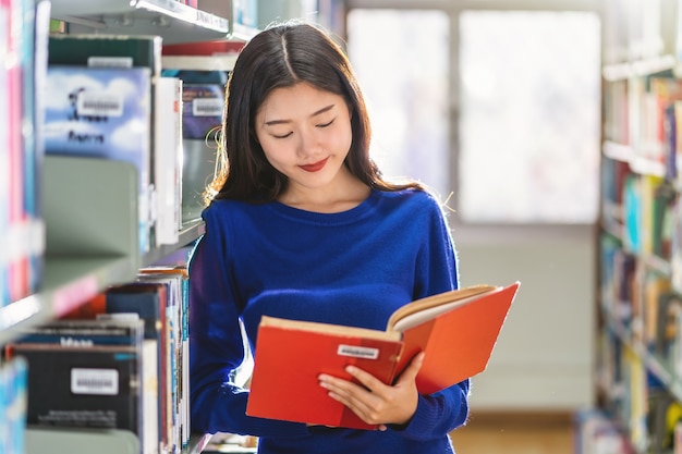 Asiatischer junger Student im Freizeitanzug, der das Buch im Bücherregal in der Bibliothek steht und liest