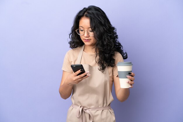 Asiatischer Kellner im Restaurant isoliert auf violettem Hintergrund, der Kaffee zum Mitnehmen und ein Handy hält