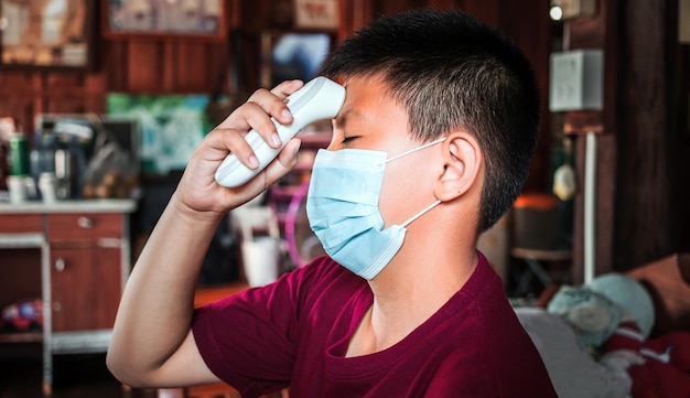 Asiatischer kinderjunge, der eine maske trägt, sieht unangenehm aus, wenn die temperatur für den fieberscan gemessen wird