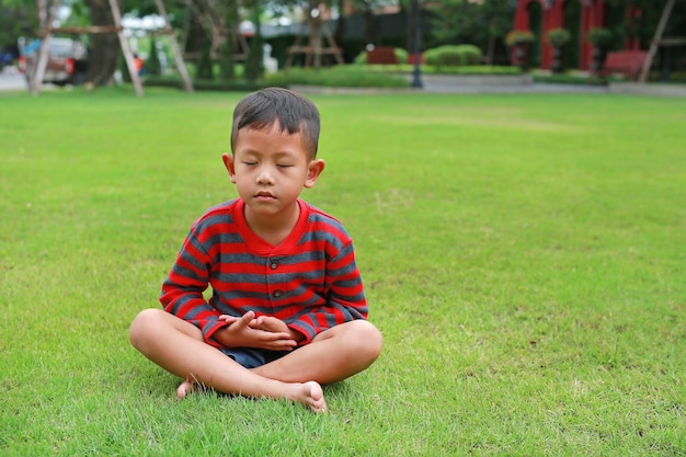 Asiatischer kleiner Junge, der Achtsamkeitsmeditation praktiziert, sitzt auf dem Rasen im Garten