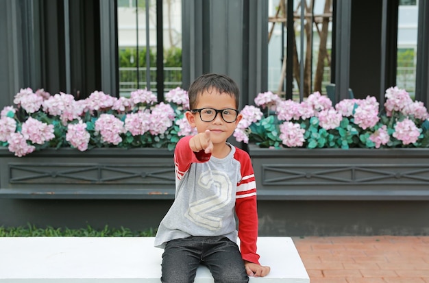 Asiatischer kleiner Junge, der auf die Kamera zeigt, während er im Garten sitzt