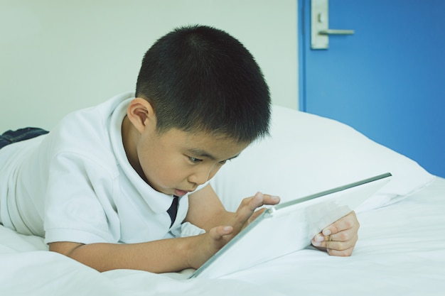 Asiatischer kleiner Junge, der Tablet-Computer im Bett verwendet
