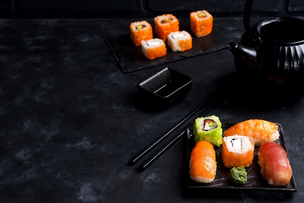 Asiatischer Lebensmittelhintergrund mit schwarzer Eisenteekanne und -sushi stellte auf Schieferplatte auf schwarzer Steintabelle ein