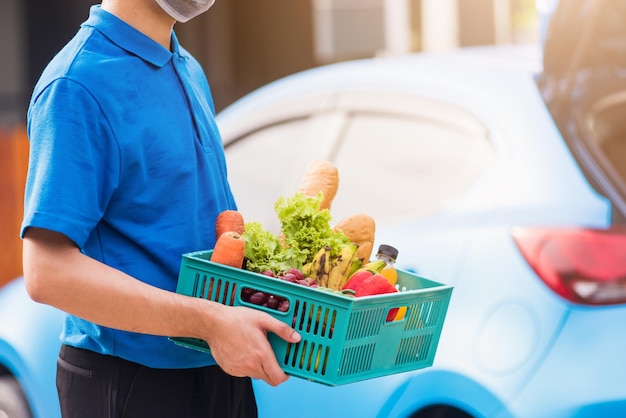 Asiatischer Lebensmittellieferant in blauer Uniform und Gesichtsmaske schützt ihn, der nach dem Ausbruch des Coronavirus frisches Gemüse in einer Plastikbox vor der Haustür liefert, zurück zum neuen normalen Konzept