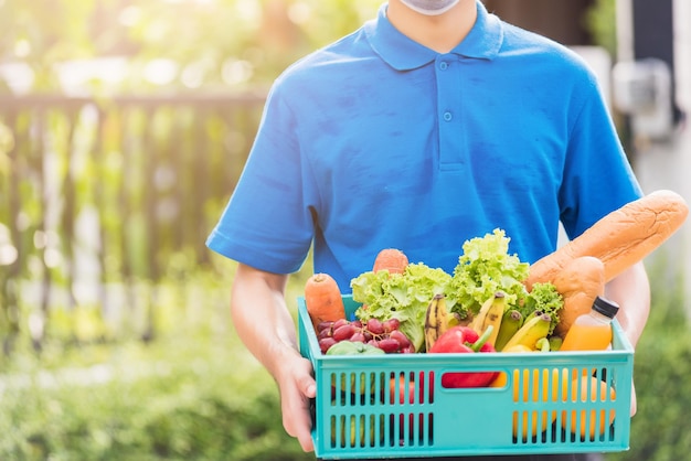 Asiatischer Lebensmittellieferant in blauer Uniform und Gesichtsmaske schützt ihn, der nach dem Ausbruch des Coronavirus frisches Gemüse in einer Plastikbox vor der Haustür liefert, zurück zum neuen normalen Konzept