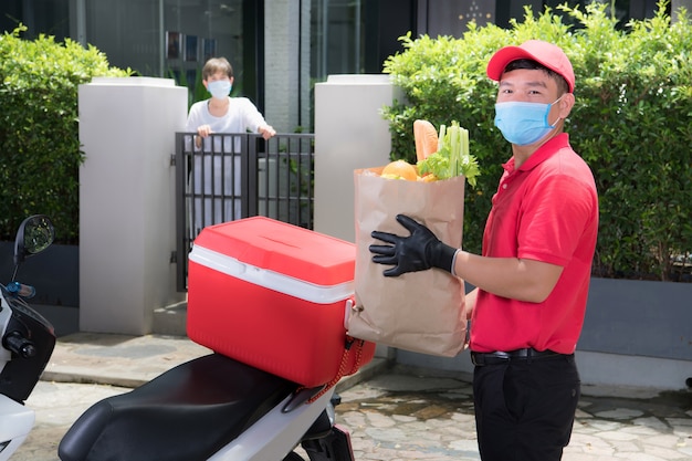 Asiatischer Lieferbote, der Gesichtsmaske und Handschuhe in der roten Uniform trägt, die Lebensmitteltasche liefert