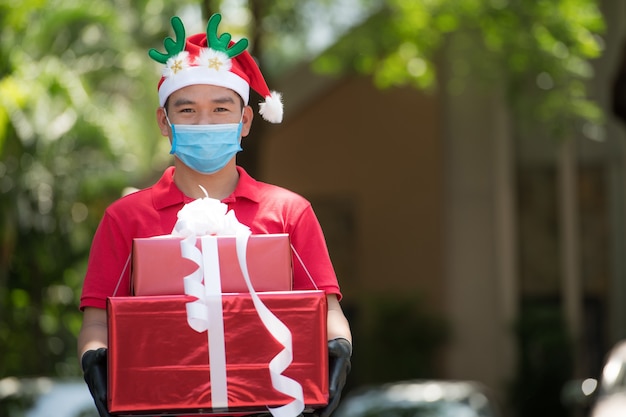 Asiatischer Lieferbote, der Gesichtsmaske und Handschuhe in roter Uniform und Weihnachtsmütze trägt, die Geschenk- und Geschenkboxen während des COVID-19-Ausbruchs für Weihnachtsfest liefern