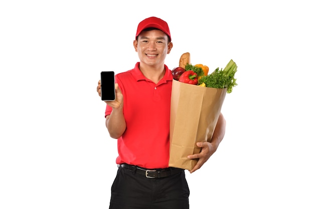 Asiatischer Lieferbote in der roten Uniform tragen Einkaufstüte und zeigt Handy lokalisiert auf weißem Hintergrund