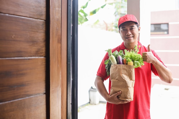 Asiatischer Lieferbote in roter Uniform, der eine Tüte mit Obst und Gemüse hält, während er den Daumen nach oben zeigt