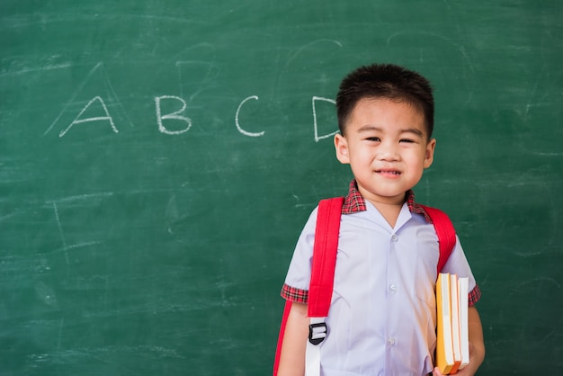 asiatischer lustiger kleiner kinderjunge aus dem kindergarten in studentenuniform mit schultasche halten bücher lächeln