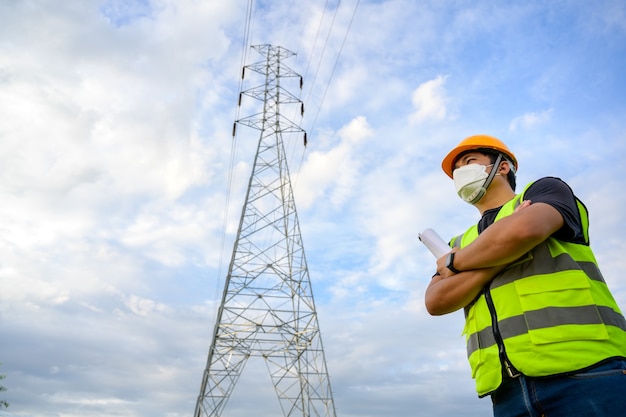 Asiatischer männlicher Elektroingenieur, der einen Bauplan im Kraftwerk hält und plant, Hochspannungsmasten im Kraftwerk zu inspizieren