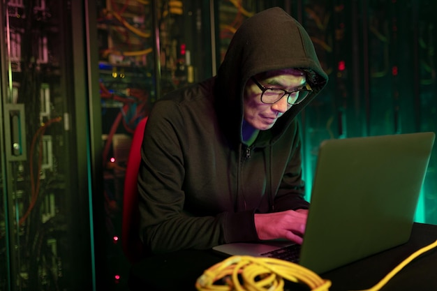 Asiatischer männlicher Hacker benutzt einen Laptop im Computerserverraum. Cyberkriminalität und Hacking-Konzept