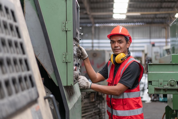 Asiatischer männlicher Ingenieur in roter Sicherheitsweste und Helm, der alte CNC-Maschinen in der Fabrik Industrial überprüft und repariert