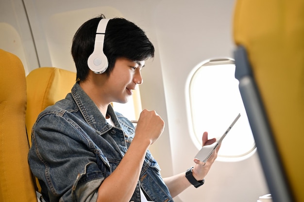 Asiatischer männlicher Passagier, der während eines Fluges mit seinem Smartphone Musik über seinen Kopfhörer hört