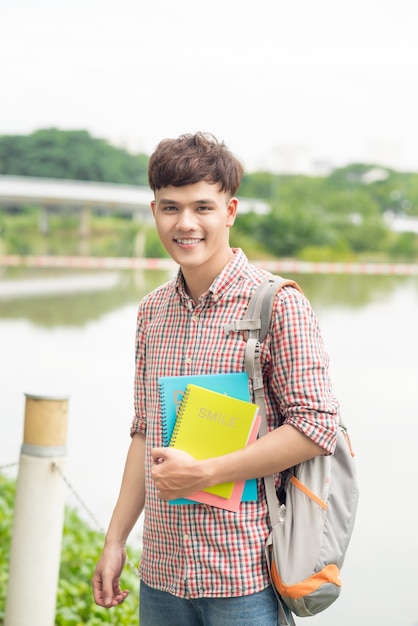 Asiatischer männlicher Student des Colleges, der Buch über den Park hält