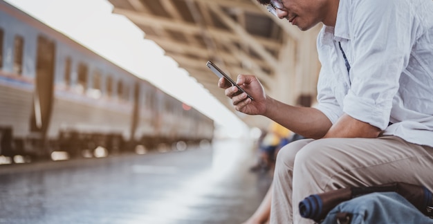 Asiatischer Mann, der am Bahnhof die richtige Richtung sucht. Reisender wartet auf den Zug und plant Ferien mit Smartphone. Urlaub, Reise, Reise und Sommer Reisekonzept.