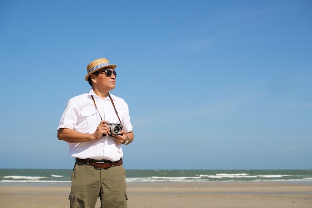 Asiatischer Mann, der auf der Sommerzeit des Strandes und des blauen Himmels mit Weinlesekamera glücklich sich fühlt