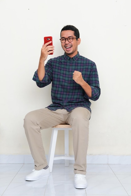 Asiatischer Mann, der auf einer Bank sitzt und Aufregung zeigt, wenn er auf sein Handy schaut