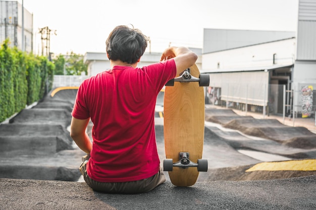 Asiatischer Mann, der bei Sonnenaufgang mit Surfskate oder Skateboard im Pumptrack-Skatepark sitzt