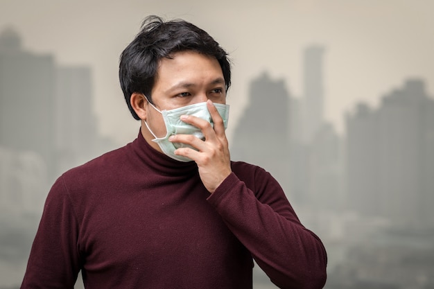 Asiatischer Mann, der die Gesichtsmaske gegen Luftverschmutzung am Balkon der hohen Wohnung trägt