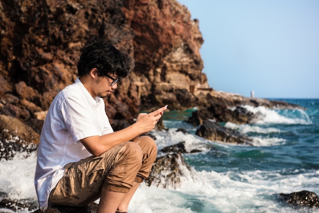 Asiatischer Mann, der die Mitteilung des Telefons schreibt, um im Sozialen Netz beim Sitzen auf Felsen auf Meer zu plaudern.