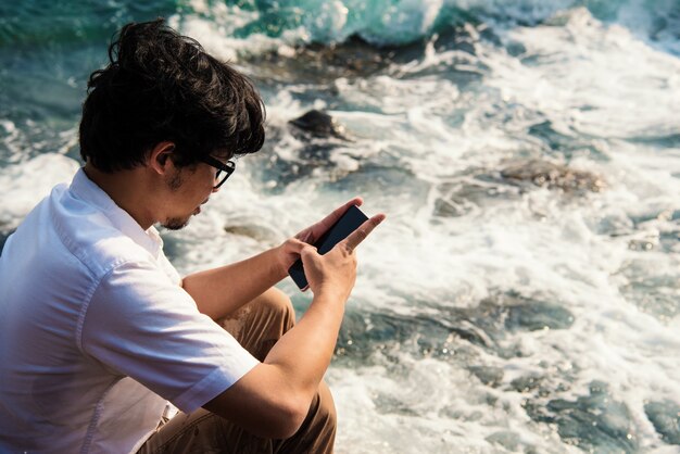 Asiatischer Mann, der die Mitteilung des Telefons schreibt, um im Sozialen Netz beim Sitzen auf Felsen auf Meer zu plaudern