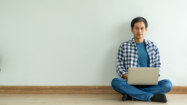 Asiatischer Mann, der ein blaues kariertes Hemd trägt, der an einem Laptop arbeitet und sich auf dem Boden des Raumes im Haus entspannt. Asiatischer Mann, Konzept der Arbeit von zu Hause aus