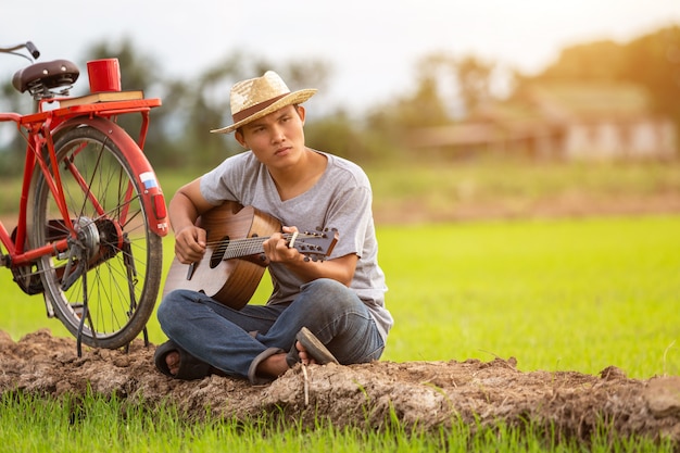 Asiatischer Mann, der Gitarre am grünen Reisfeld in der Sonnenuntergangzeit spielt