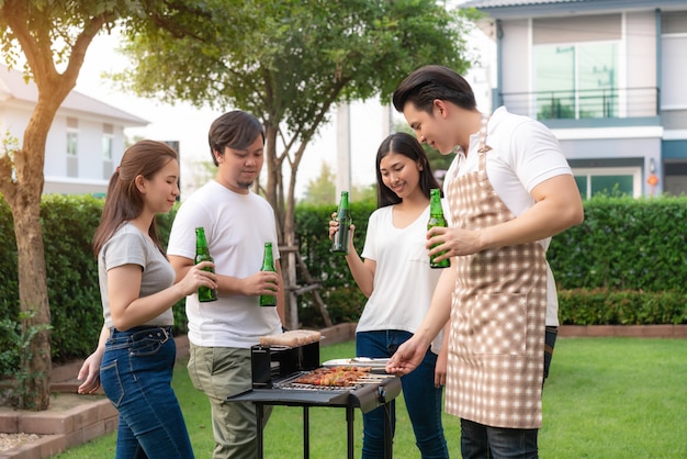 Asiatischer Mann, der Grill und Wurst kocht, damit eine Gruppe von Freunden Party im Garten zu Hause isst.