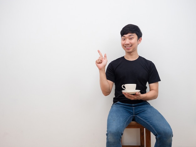 Asiatischer Mann, der Kaffeetasse hält und mit dem Finger auf den Raum zeigt, mit einem glücklichen Lächeln, das auf einem Stuhl auf weißem Hintergrund sitzt