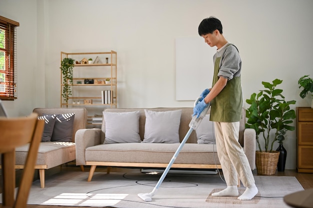 Asiatischer Mann, der sich darauf konzentriert, sein Wohnzimmer mit einem Staubsauger zu reinigen, um den Teppich zu reinigen