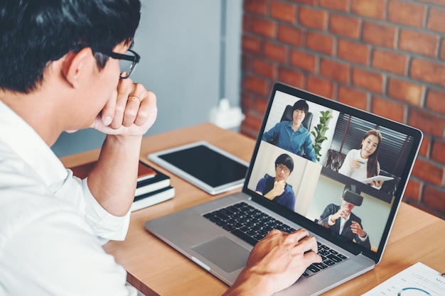 Asiatischer Mann, der von zu Hause aus arbeitet, nutzt ein Online-Meeting für intelligentes Arbeiten und Videokonferenzen mit einem asiatischen Team, das Laptop und Tablet online im Videoanruf für neue Projekte verwendet