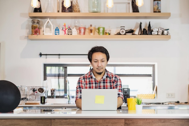 Asiatischer Mann, der von zu Hause aus mit Technologie-Laptop und Mobiltelefon im Küchenraum arbeitet