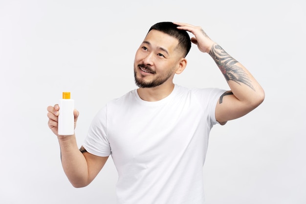 Asiatischer Mann hält Flasche mit Haarshampoo und schaut isoliert auf weißem Hintergrund weg. Haarpflege