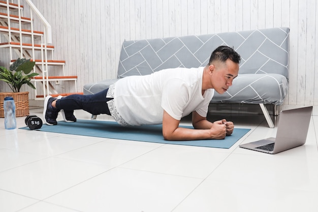 Asiatischer Mann im weißen Hemd auf blauer Matte, der Planke macht, während er eine Video-Fitnessstunde auf dem Laptop sieht