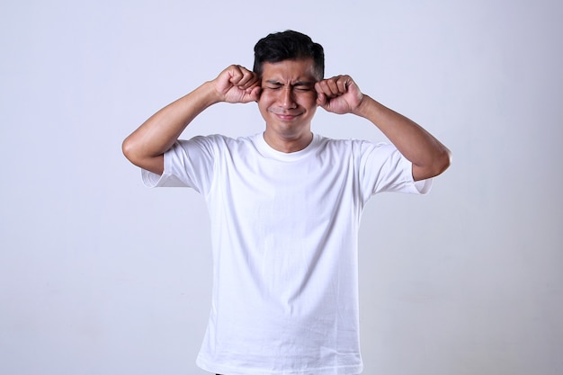 Asiatischer Mann im weißen T-Shirt mit lustigem weinenden Ausdruck auf weißem Hintergrund