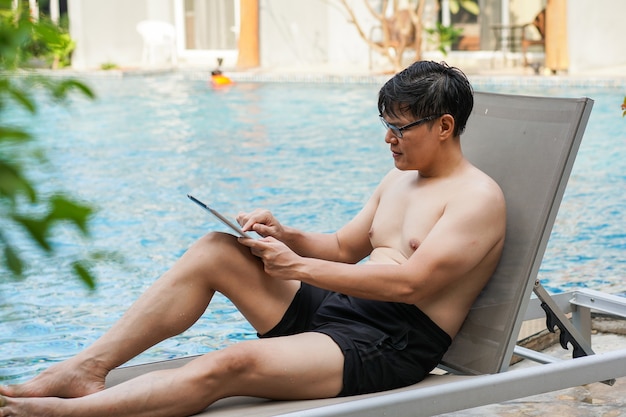 Asiatischer Mann in Badeshorts, der auf einem digitalen Tablet beim Schwimmen spielt