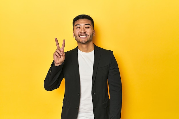 Asiatischer Mann in schwarzer Geschäftsjacke auf einem gelben Studio zeigt ein Siegeszeichen und lächelt breit
