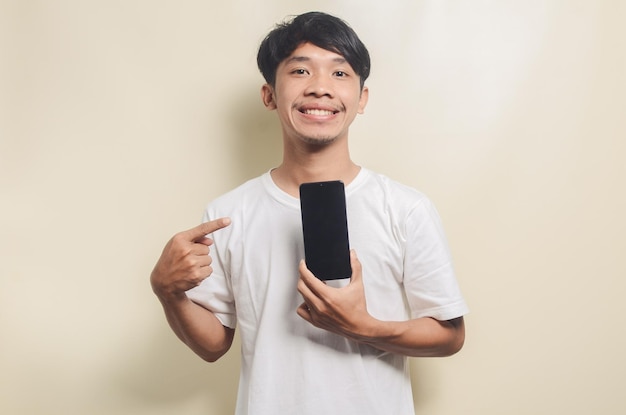 Asiatischer Mann in weißem T-Shirt mit Geste, der sein Telefon auf isoliertem Hintergrund zeigt