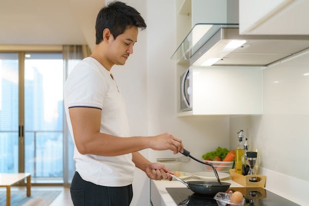 Asiatischer Mann kocht amerikanisches Frühstück, indem er ein Spiegelei in der Pfanne in ihrer Küche zu Hause brät.